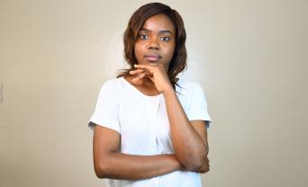 A young woman wearing a white shirt stands against a plain light-colored wall, resting one arm across her body and her chin on her hand in a thoughtful pose. She looks directly at the camera with a calm, serious expression.