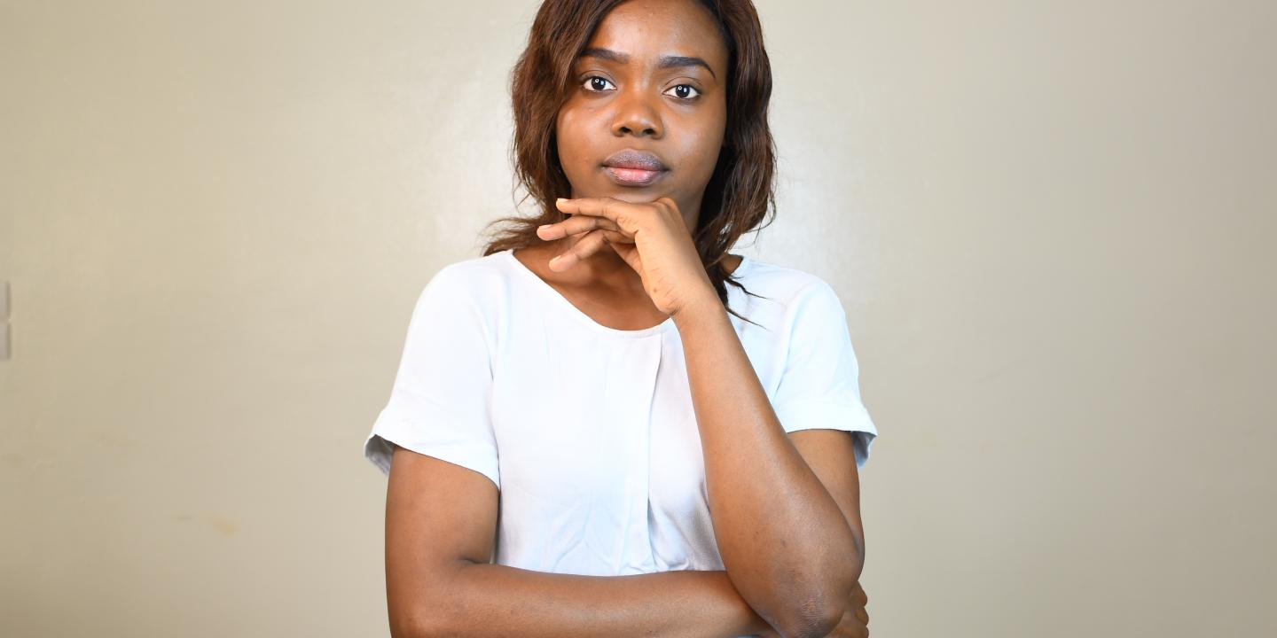 A young woman wearing a white shirt stands against a plain light-colored wall, resting one arm across her body and her chin on her hand in a thoughtful pose. She looks directly at the camera with a calm, serious expression.